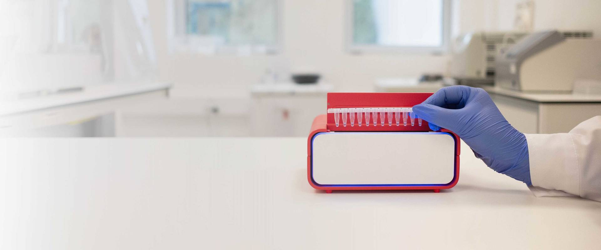 The InviDx® instrument positioned on a laboratory bench, with a gloved hand placing a strip of sample tubes into the device. The bright red and white instrument is shown in a clean, modern laboratory setting with blurred laboratory equipment in the background.