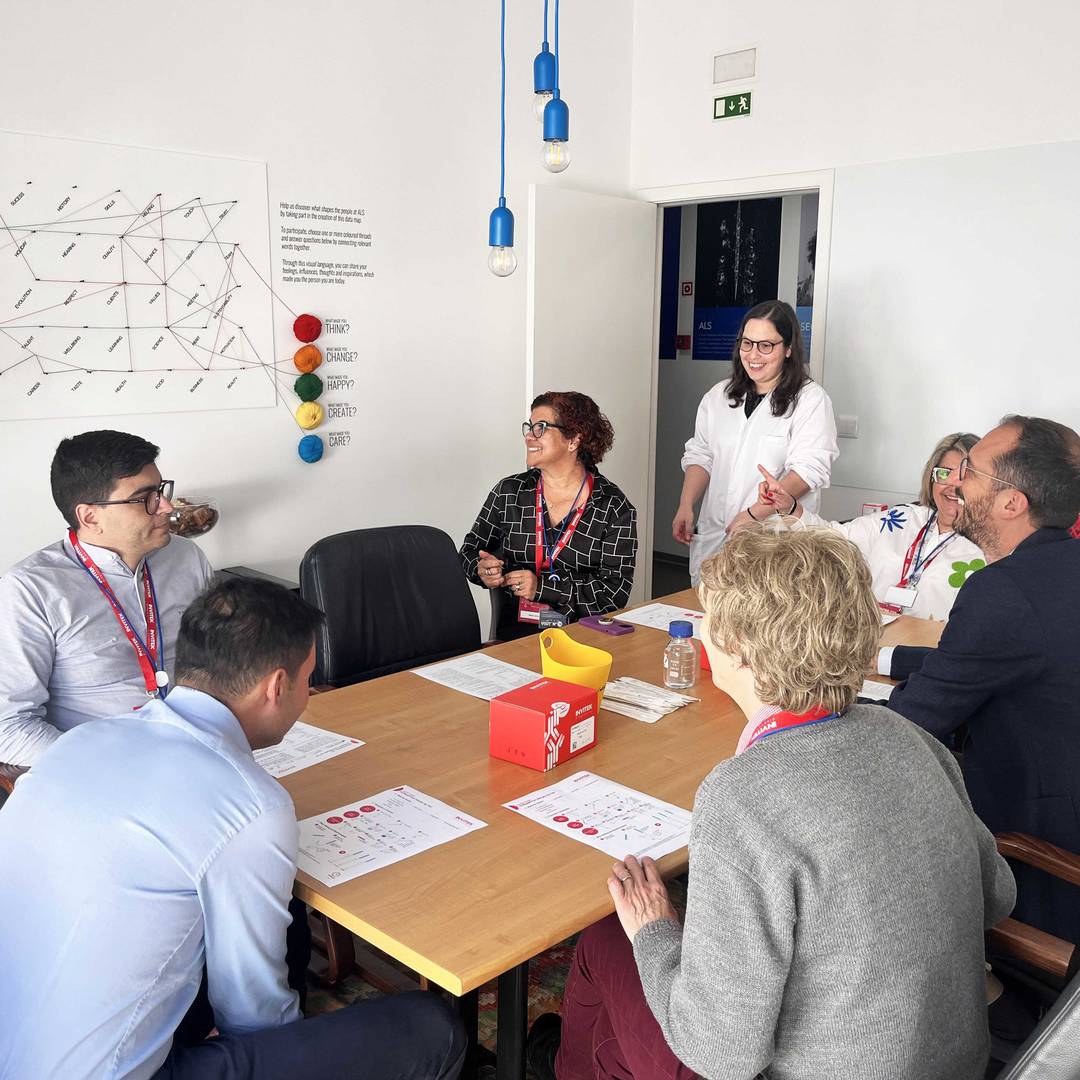 Group of seven people sitting around a table with papers and a red box, engaged in discussion or activity, with a chart and colored balls on the wall behind them.