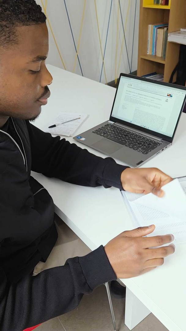 A man sits at a white desk, reading papers beside an open laptop in a tidy study area with shelves and books in the background