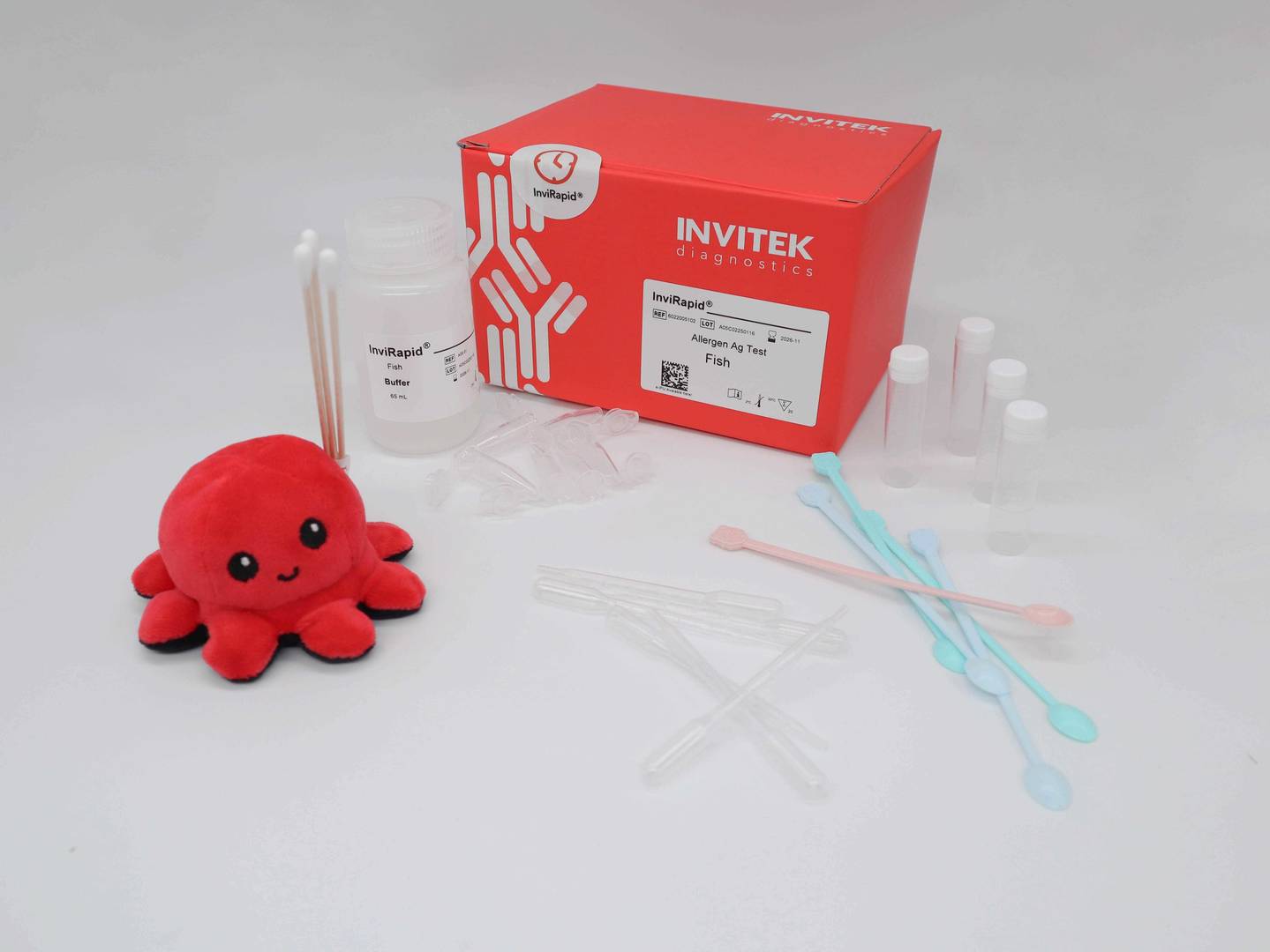Red Invitek Diagnostics InviRapid Fish Allergen Ag Test box behind a buffer bottle and sample tubes next to swabs with scoops, droppers and tubes laying scattered in front.