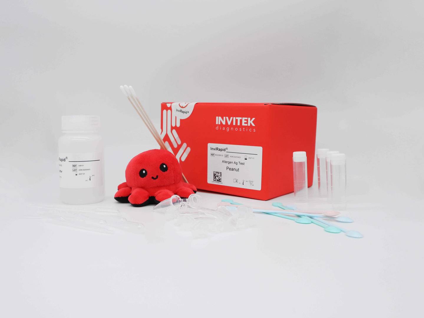Red Invitek Diagnostics InviRapid Peanut Allergen Ag Test box behind sample tubes and swabs next to a buffer bottle with scoops, droppers and tubes laying scattered in front.