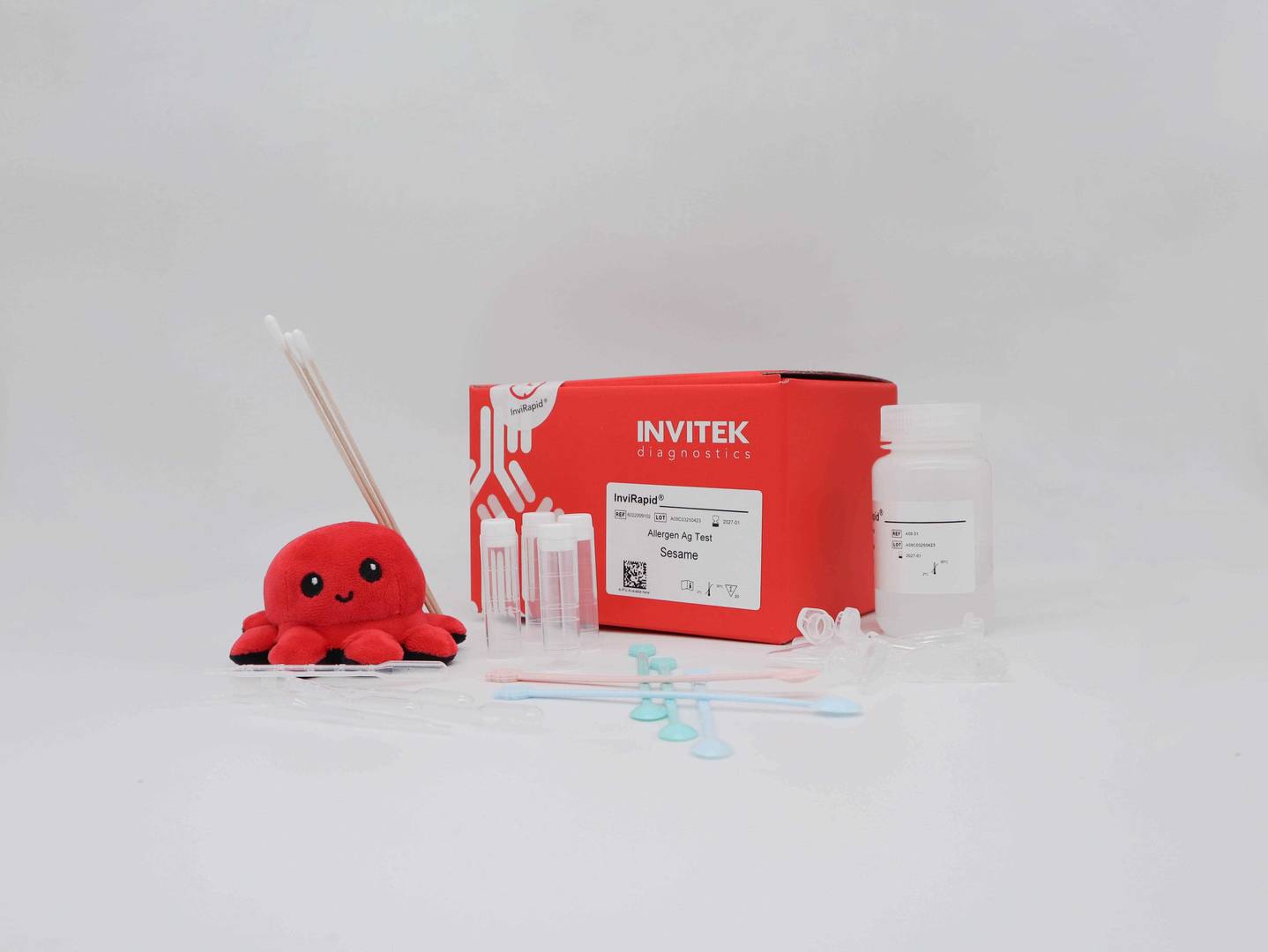 Red Invitek Diagnostics InviRapid Sesame Allergen Ag Test box behind a buffer bottle and sample tubes next to swabs with scoops, droppers and tubes laying scattered in front.