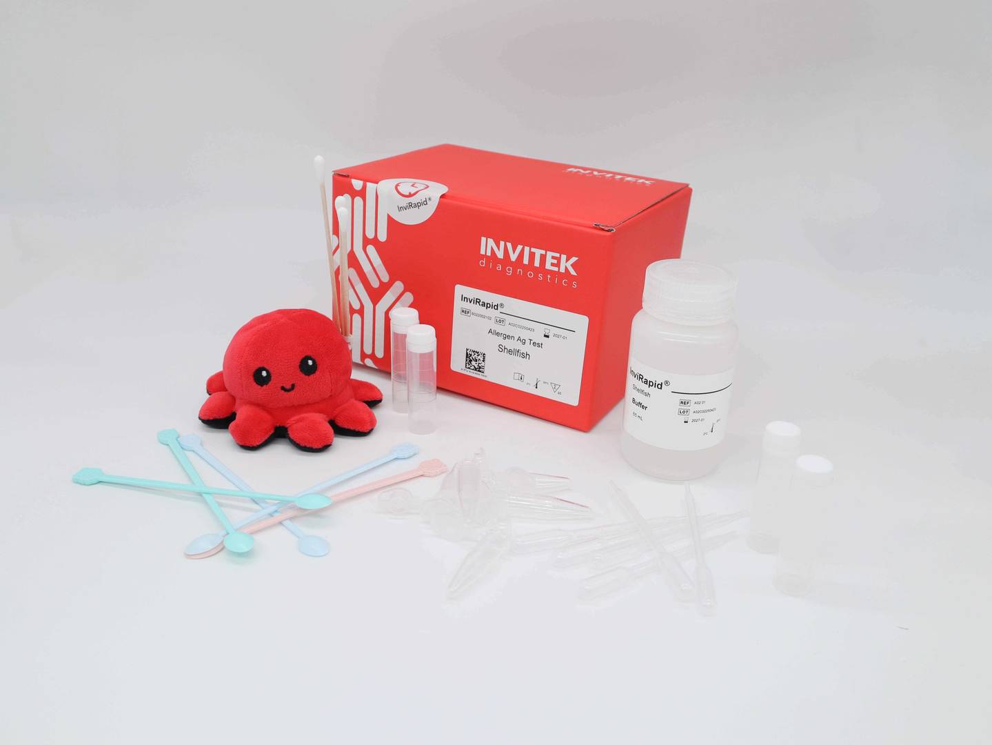 Red Invitek Diagnostics InviRapid Shellfish Allergen Ag Test box behind a buffer bottle, sample tubes, and swabs with scoops, droppers and tubes laying scattered in front.
