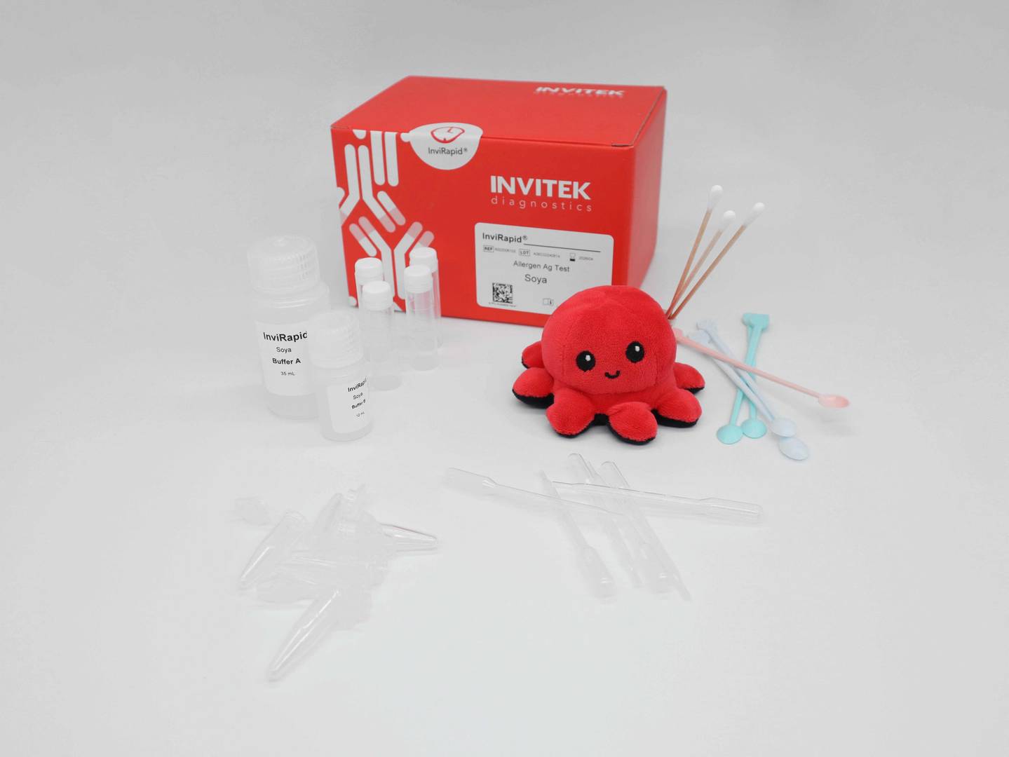 Red Invitek Diagnostics InviRapid Soya Allergen Ag Test box behind buffer bottles, sample tubes, and swabs with scoops, droppers and tubes laying scattered in front.