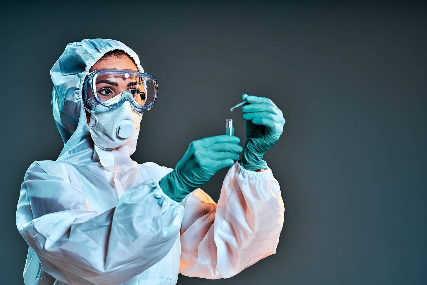 Person wearing a full-body cleanroom suit, protective goggles, respirator mask, and green gloves carefully handling a small vial and pipette in a controlled laboratory environment.
