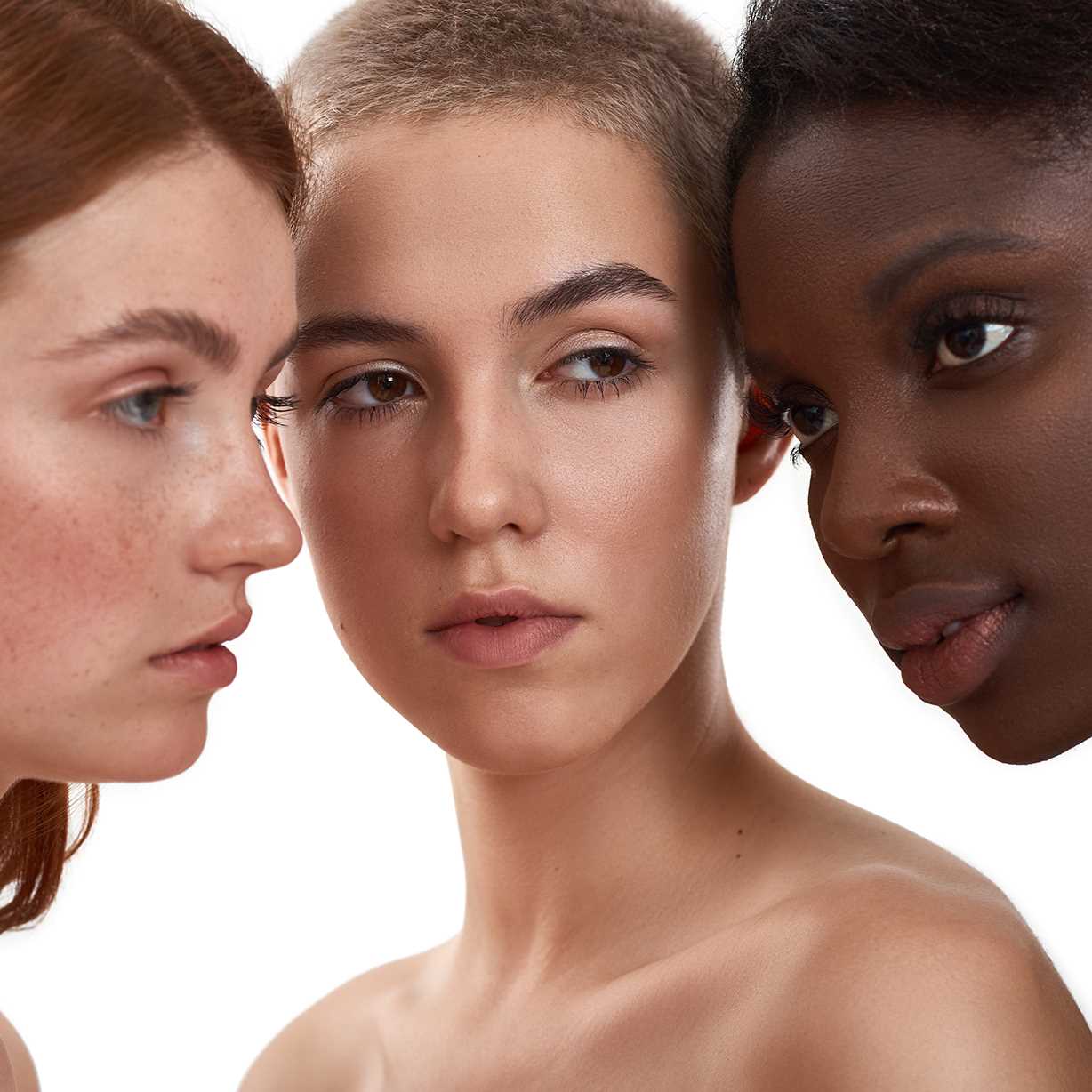 Close-up portrait of three women with different skin tones positioned face-to-face against a white background.